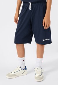 Kind in marineblauwe Champion-shorts, witte Champion-sokken en witte sneakers met blauwe accenten, staande voor een effen achtergrond.
