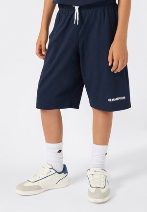 Enfant portant un short Champion bleu marine, des chaussettes Champion blanches et des baskets blanches avec des accents bleus, se tenant devant un fond uni.