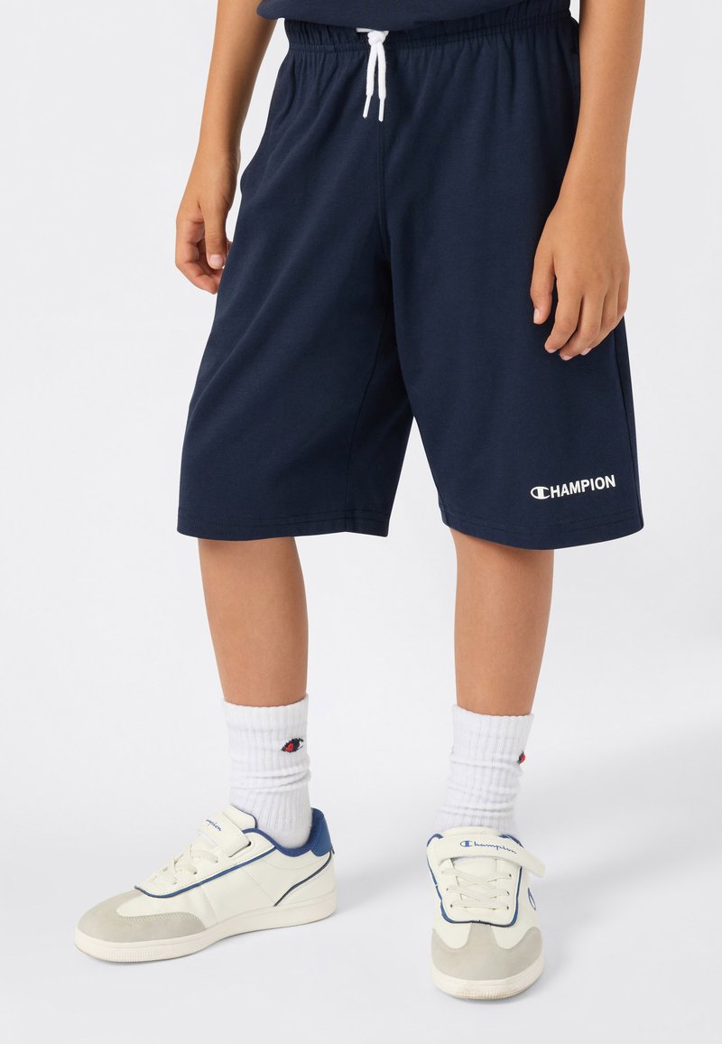 Kind in marineblauwe Champion-shorts, witte Champion-sokken en witte sneakers met blauwe accenten, staande voor een effen achtergrond.