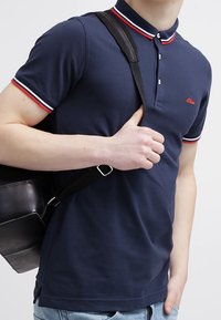 Granatowa koszulka polo z kołnierzykiem w czerwono-białe paski, krótkie rękawy, zapięcie na trzy guziki i małym logo na klatce piersiowej.