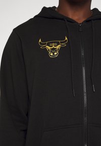 Sudadera con cremallera negra hecha de tela suave, con un logo de toro dorado en el lado izquierdo del pecho, capucha con cordón y bolsillos frontales.