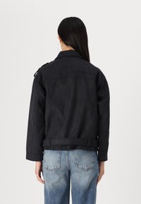 Black suede jacket with a structured collar, side pockets, and a fitted waist. Paired with faded blue jeans, showcasing a relaxed fit.