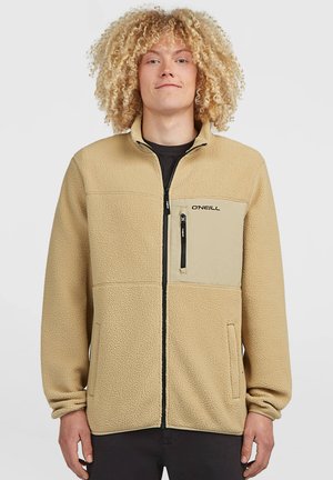 Junge Person mit lockigem blondem Haar, die eine beige Fleecejacke mit einer Reißverschlusstasche auf der Brust trägt und vor einem schlichten Hintergrund steht.
