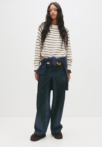Gestreifter cremefarbener und brauner Pullover, dunkelblauer Sweatshirt um die Taille gebunden, weit geschnittene dunkle Jeans und braune Slipper.