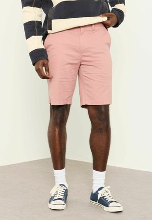 Person trägt hellrosa knielange Shorts, ein langärmeliges Shirt mit marineblauen und cremefarbenen Streifen, weiße Socken und navyblaue Canvas-Sneaker.