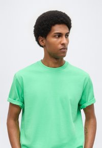 Νέος άνδρας με κοντά σγουρά μαλλιά φοράει φωτεινό πράσινο κοντομάνικο μπλουζάκι με γυρισμένα μανίκια, κοιτάζει στο πλάι με φόντο απλό φωτεινό.