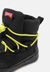 Bota negra con parte superior texturizada, cordones elásticos amarillos y un logo de Camper. Presenta una suela negra moteada y un collar acolchado para mayor soporte.