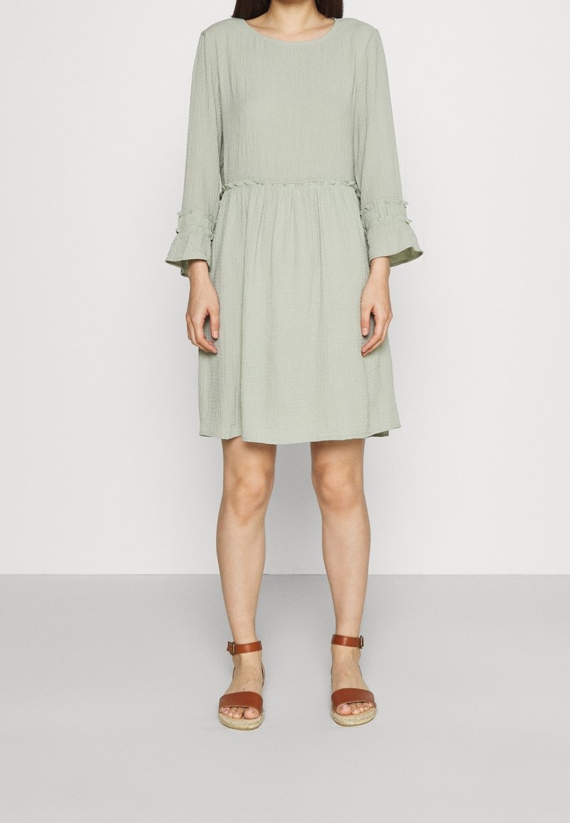 Femme portant une robe vert clair, longueur genou avec manches longues et sandales marron à bout ouvert avec bride à la cheville, debout devant un fond uni.