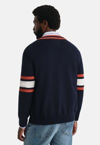 Cardigan tricotat navy cu accente rayate în alb, roșu și negru pe manșete. Prezintă un tiv cu nervuri și o croială relaxată.