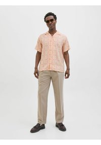 Kurzarmhemd in Pastellrosa mit geometrischem Muster, kombiniert mit beigen Hosen und dunklen Schuhen. Das Modell trägt außerdem Sonnenbrillen.