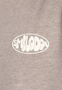 Grå bomuldsstof har et tekstureret logo i hvidt med rundede, flydende bogstaver. Designet er centreret inden for en oval form.