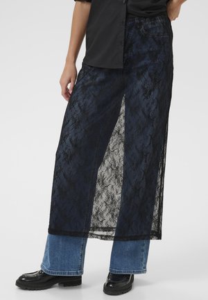 Gonna lunga e stratificata con un sottile strato di pizzo nero trasparente sopra il denim scuro, con un orlo appena sopra le caviglie e jeans blu a contrasto sotto.
