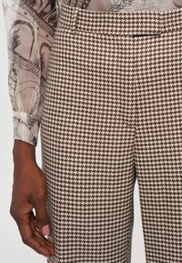 Bruine en crème houndstooth-gepatenteerde broek met een rechte pasvorm, voorzien van een op maat gemaakte tailleband en een gladde textuur. Hand getoond.