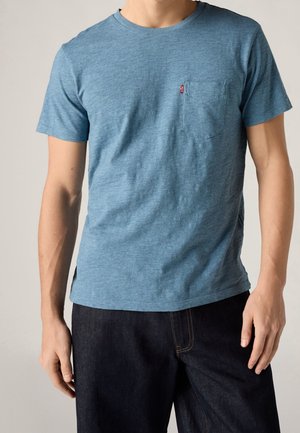 Homme portant un t-shirt bleu à manches courtes, col rond avec une poche poitrine et un jean en denim foncé sur un fond uni.