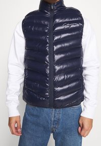 Man som bär en glänsande marinblå kviltad ärmlös väst över en vit långärmad skjorta och blå jeans, står med avslappnade händer.