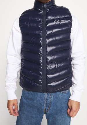 Un uomo indossa un gilet trapuntato senza maniche blu navy lucido sopra una camicia bianca a maniche lunghe e jeans blu, in piedi con le mani rilassate.