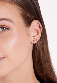 Créole en or avec des accents noirs, trois clous d'oreilles en argent et en or, comprenant une perle, une étoile et une pierre claire. Cheveux bruns et raides.