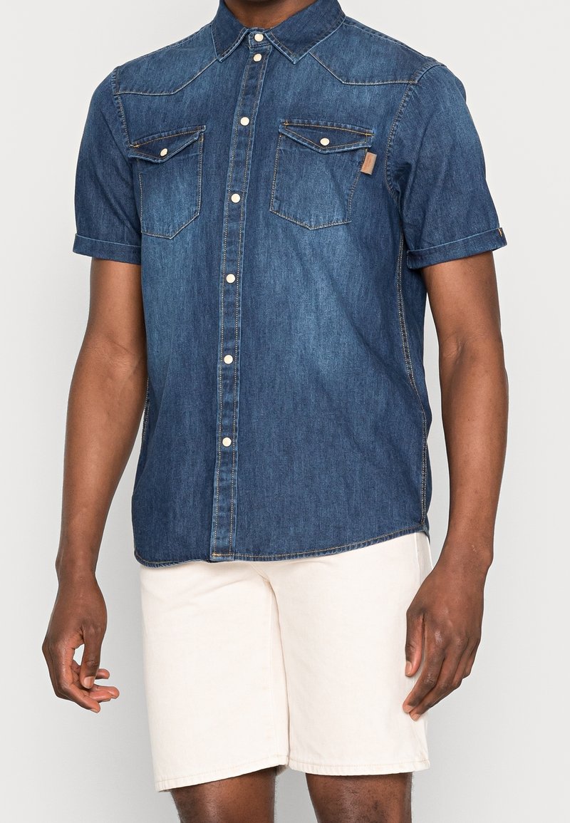 Homme portant une chemise en denim à manches courtes bleu foncé à boutons et un short beige clair, debout devant un fond blanc uni.