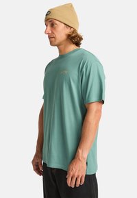 Camiseta de manga corta verde menta con cuello redondo, que presenta un pequeño logo dorado en el pecho, combinada con un gorro beige.