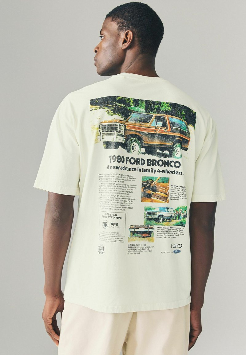 Tricou din bumbac de culoare bej deschis, cu un grafic mare retro al Ford Bronco pe spate, având text și imagini colorate legate de vehicul.