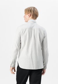 Camicia a righe grigia chiaro e bianca, con maniche lunghe, polsini abbottonati e dettaglio della yoke sul retro. Materiale in cotone dalla texture morbida.