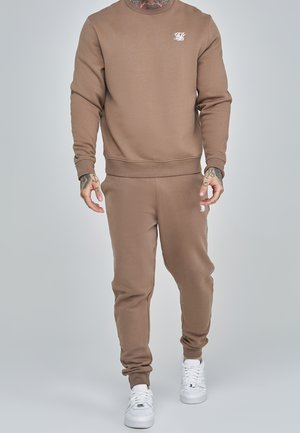 Mann trägt einen braunen Sweatshirt und passende Jogginghose mit weißen Sneakers und sichtbaren Tattoos an Händen und Hals.