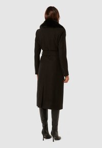 Abrigo de lana negro con cuello de piel sintética, cintura ceñida con cinturón, mangas largas y detalle de abertura en la parte trasera. Combinado con botas negras de tacón hasta la rodilla.
