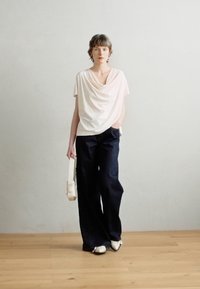 Svetlerůžový top s volně splývavým střihem, tmavé široké kalhoty, bílé boty. Modelka drží bílou tašku, stojí na dřevěné podlaze na pozadí s jednobarevnou plochou.