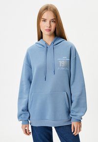 Sudadera azul claro con bolsillo canguro, puños acanalados y un estampado gráfico con "1991" y "Los Ángeles". Tela suave y texturizada.