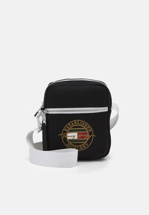 Sac bandoulière carré noir avec bandoulière blanche réglable, poche avant zippée et écusson rond avec logo brodé doré.