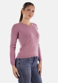 Maglione rosa a V con maniche lunghe, design aderente e orlo a costine. Indossato con jeans blu scuro, tessuto testurizzato e dettagli minimi.