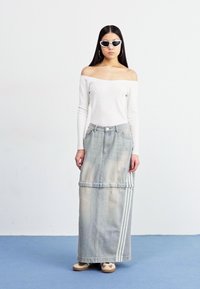 Jupe maxi en denim bleu délavé avec deux rayures latérales, associée à un haut ajusté à manches longues blanc cassé et des lunettes de soleil rondes. Chaussures plates.