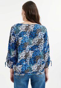 Blusa floreale blu, nera e bianca con maniche a tre quarti, un orlo leggermente arricciato e un'apertura a chiave sul retro con un bottone.