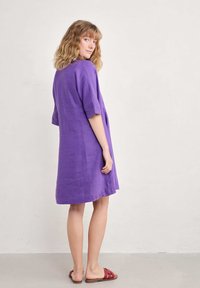 Violet linen dress with short sleeves, knee-length cut, relaxed fit, and side pockets. Paired with red slide sandals on a neutral background.