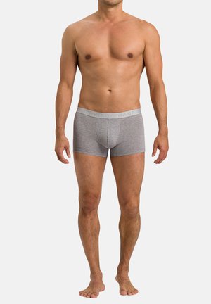 Man staat op blote voeten en draagt grijze boxershorts met een gebrandmerkte tailleband tegen een effen lichte achtergrond.
