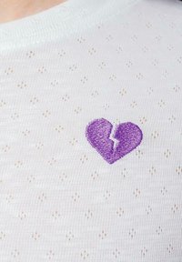 Tejido de punto blanco con un patrón texturizado, con un diseño de corazón roto bordado en púrpura en el centro.