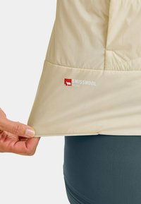 Beige Jacke aus einem glatten, leichten Stoff. Verfügt über eine Swisswool-Markierung am unteren Saum. Die Hand greift den Stoff am Saum.