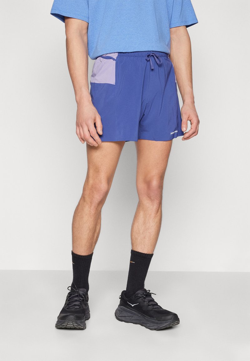 Calções de desporto azuis com um bolso de destaque roxo, cintura elástica e cordão. Usados com sapatos e meias pretas, em pé contra um fundo branco.