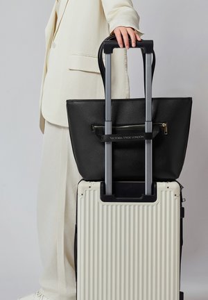 Persona con traje crema sosteniendo un bolso negro Victoria Hyde London, sujeto al mango extendido de una maleta rígida blanca con relieve acanalado.