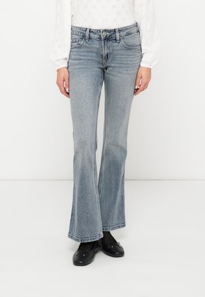 Person trägt hellblaue ausgestellte Jeans, weiße Langarmbluse mit Lochstickerei, schwarze Strumpfhosen und schwarze Ballerinas und steht.