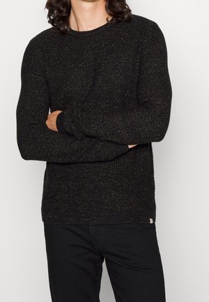 Pull en maille noir foncé avec des éclats dorés, col rond, design ajusté et poignets côtelés, porté avec un pantalon sombre.