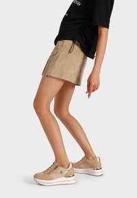 Sneakers beige con tessuto testurizzato e accenti lisci, dotati di una suola a piattaforma stratificata e dettagli dorati. Indossati con una camicia nera e una gonna in suede color beige.