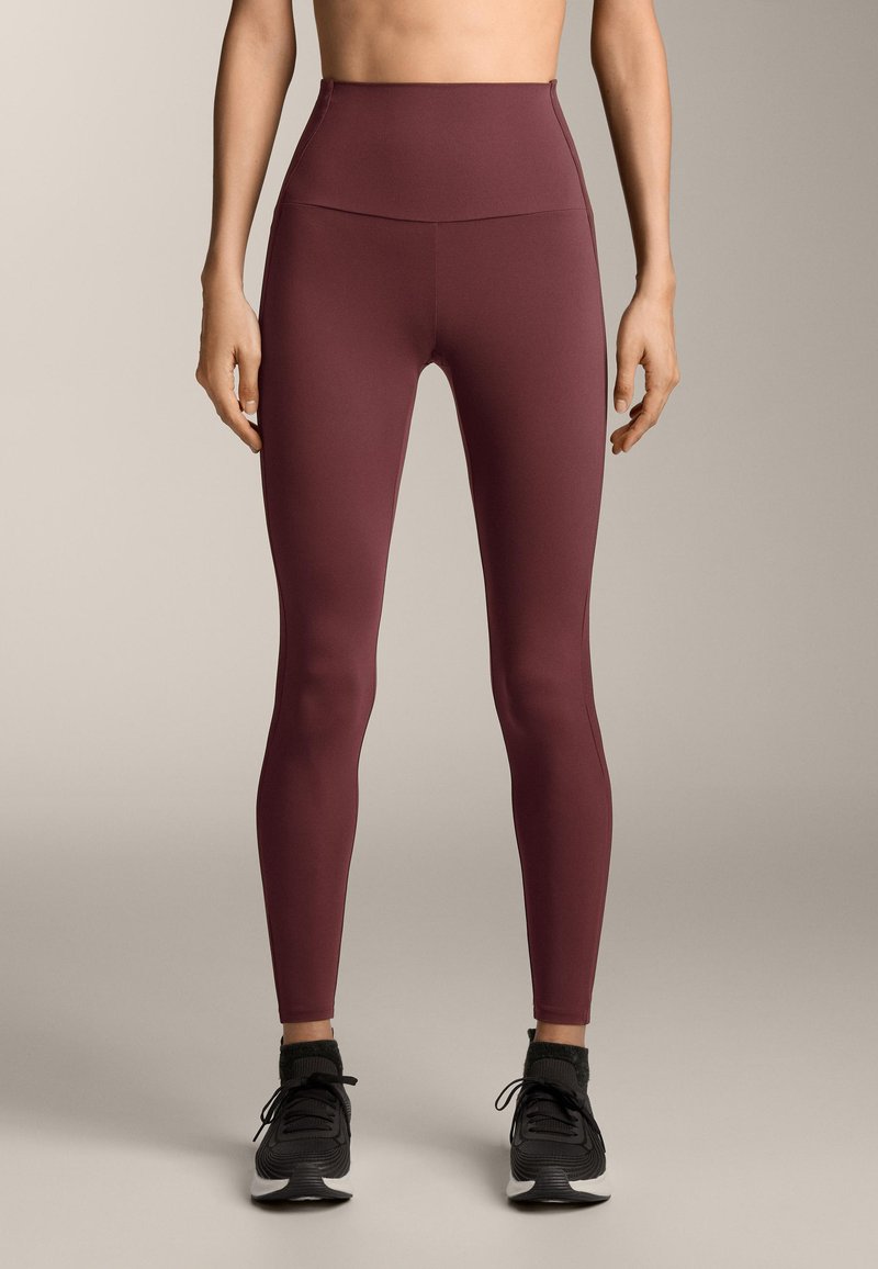 Leggings bordeaux a vita alta in materiale elastico, caratterizzati da una texture liscia e una silhouette aderente, abbinati a scarpe sportive nere.