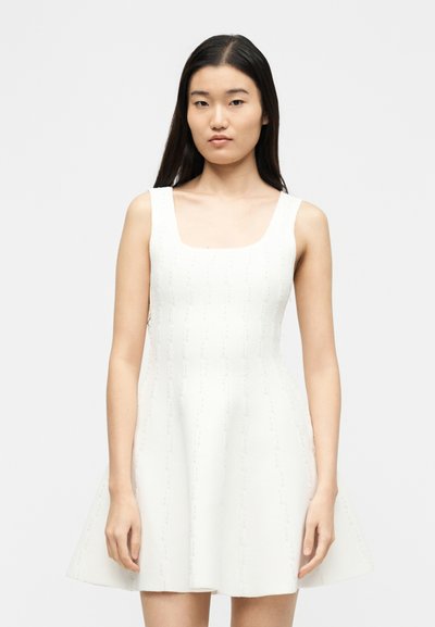 Νεαρή γυναίκα με μακριά σκούρα μαλλιά φοράει αμάνικο λευκό πλεκτό φόρεμα με υφασμένο κατακόρυφο μοτίβο από κουκκίδες, στέκεται μπροστά σε απλό ανοιχτό φόντο.