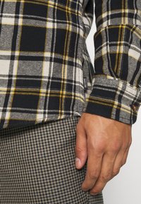 Camisa de franela de cuadros en negro y amarillo con puños abotonados, combinada con pantalones de cuadros grises. Textura suave con detalles de costura visibles.