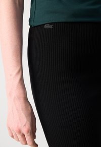 Fustă neagră cu model canelat și un design ajustat, însoțită de un top verde închis. Detaliul remarcabil include un mic accent cu logo-ul Lacoste.