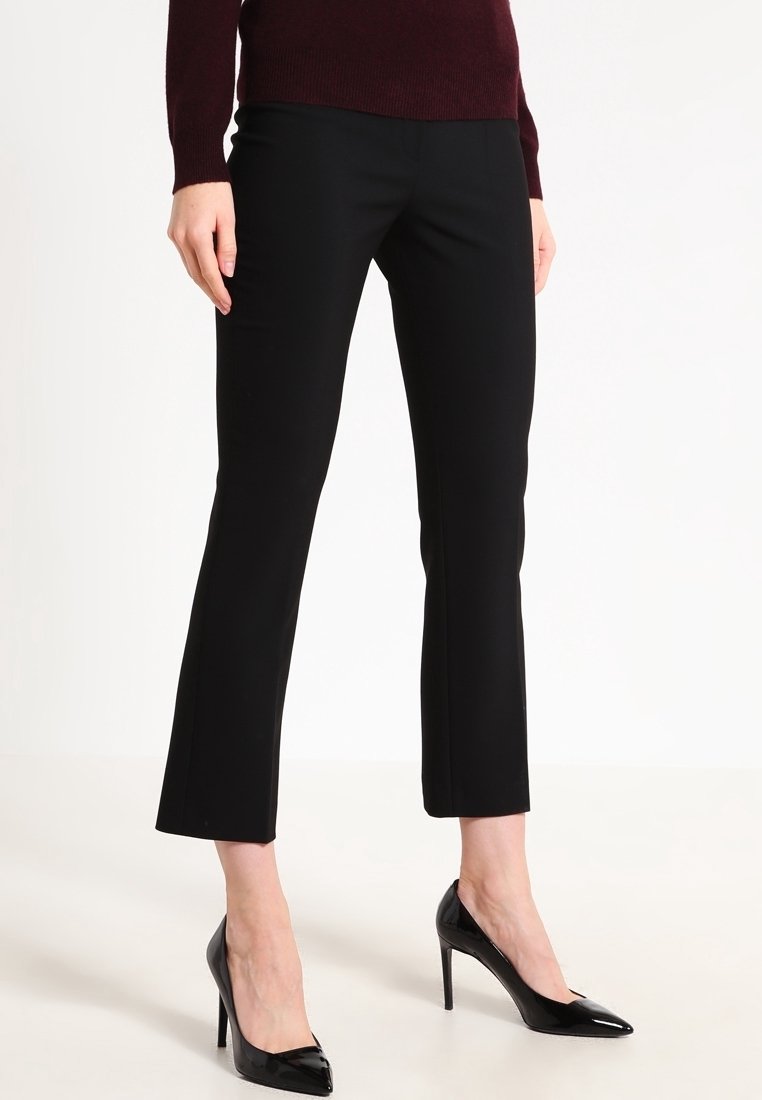 Femme portant un pantalon droit noir, un pull bordeaux et des chaussures à talons hauts en cuir verni noir, debout devant un fond blanc.