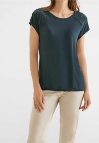 Tmavě zelený krátký rukáv s jemnou strukturou, zdobený krajkovým lemem na ramenech a volným střihem, v kombinaci se světlými béžovými kalhotami.