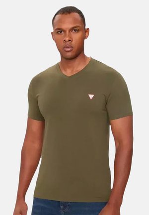 Homme portant un t-shirt vert olive à manches courtes et col en V avec un petit logo triangulaire sur la poitrine et un jean bleu, debout sur un fond uni.