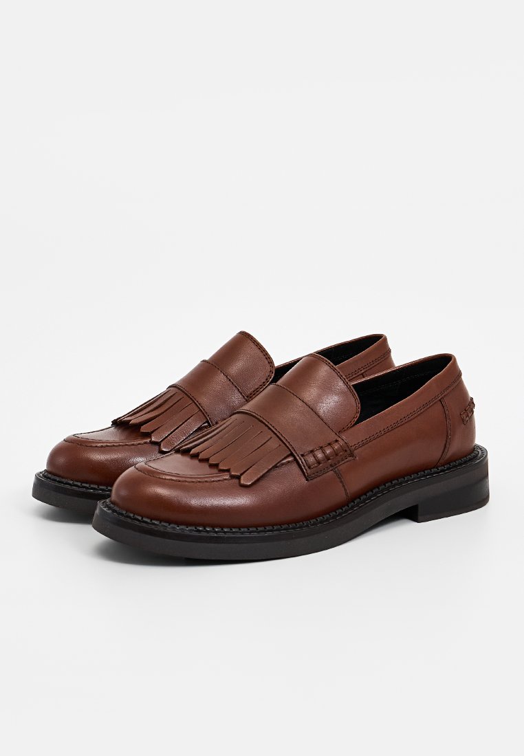 Mocasines de cuero marrón con una correa con flecos, punta redonda, suela baja negra y detalles de costura en la parte superior.